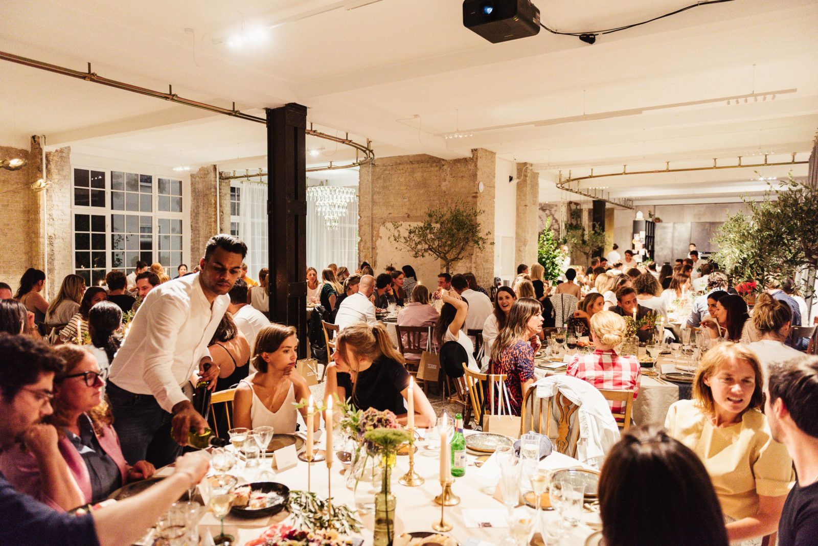 Guests seated at long tables under gold pendant lamps in the Friends Space, Berlin