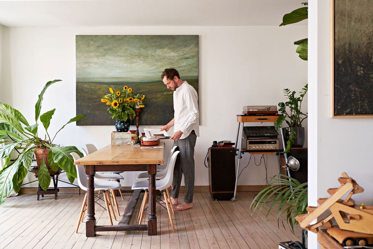 Painter at a long wooden table in his plant-filled flat in Antwerp, a landscape painting on the wall
