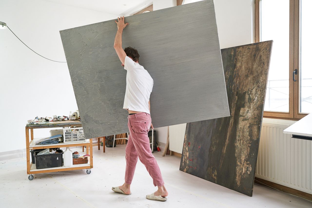 Painter carrying a large grey canvas across his white studio in Antwerp