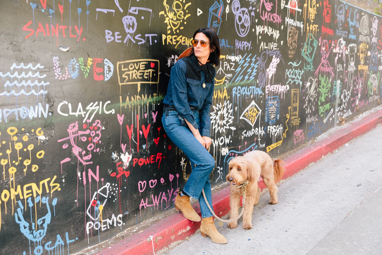 Stylist in denim leaning on a chalk-covered wall with her dog, Los Angeles