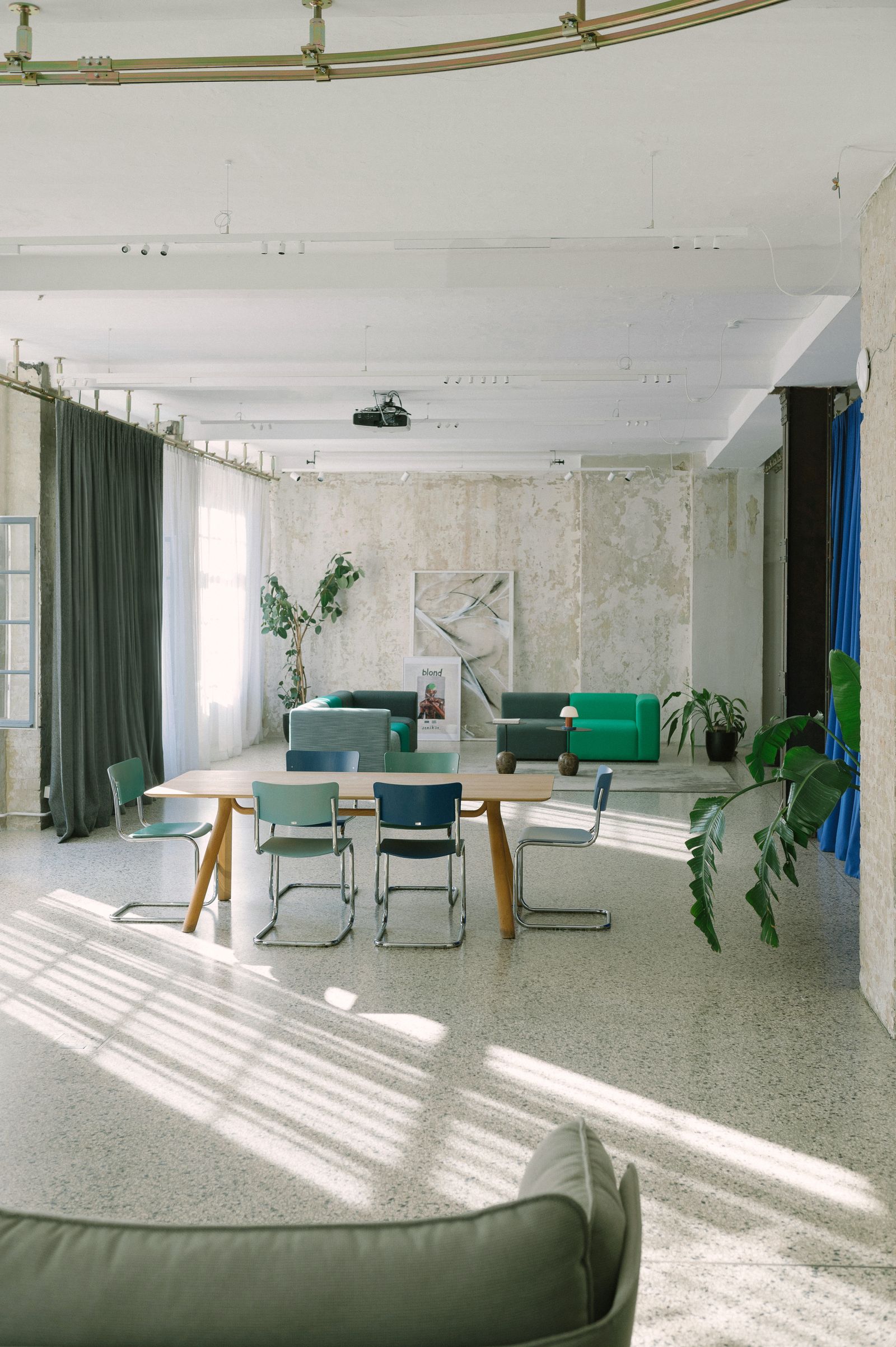 Long room with tables, steel chairs and green sofas in the Friends Space, 2024