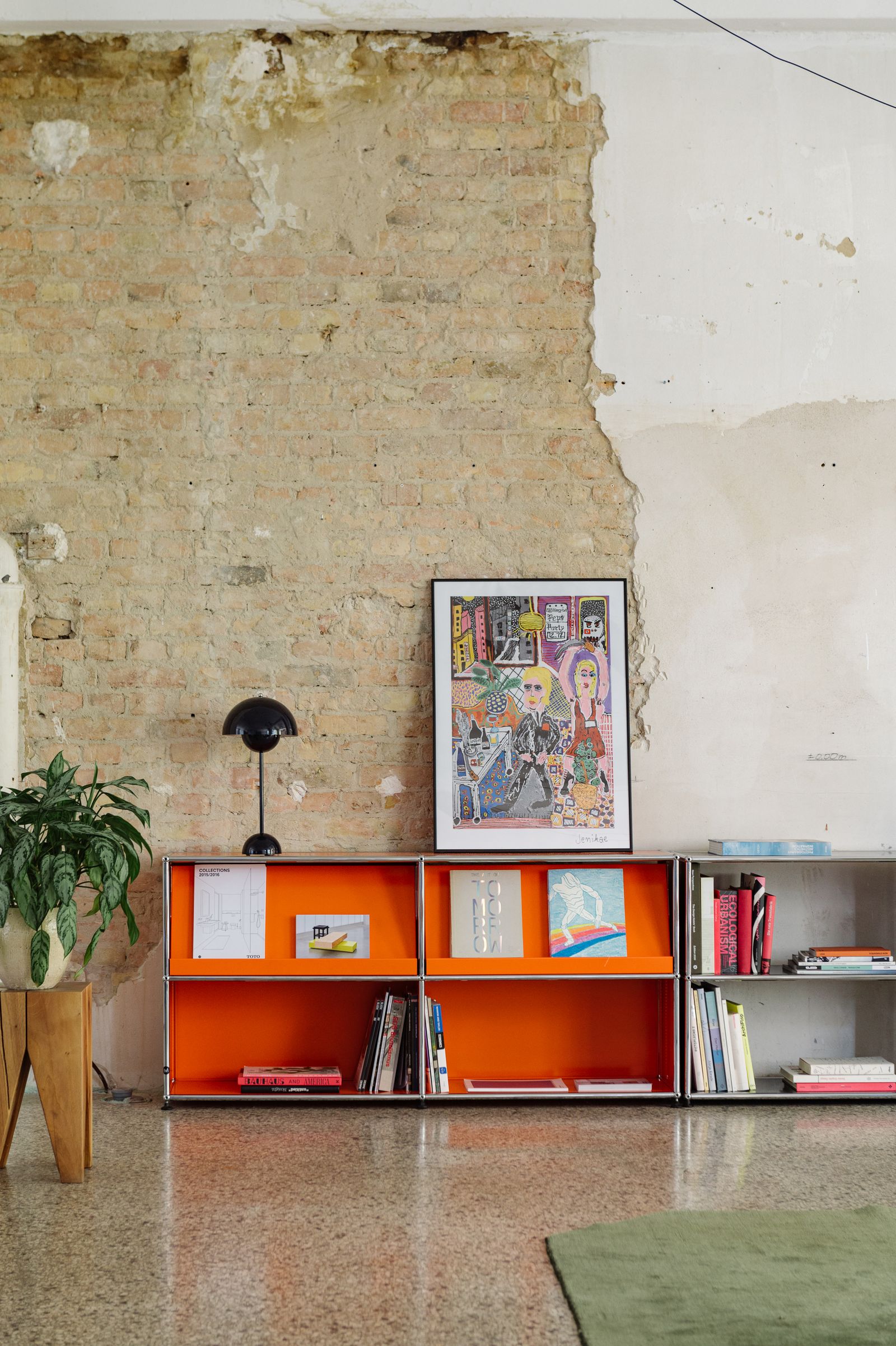 Orange shelf with books and a framed print against an exposed brick wall in the Friends Space