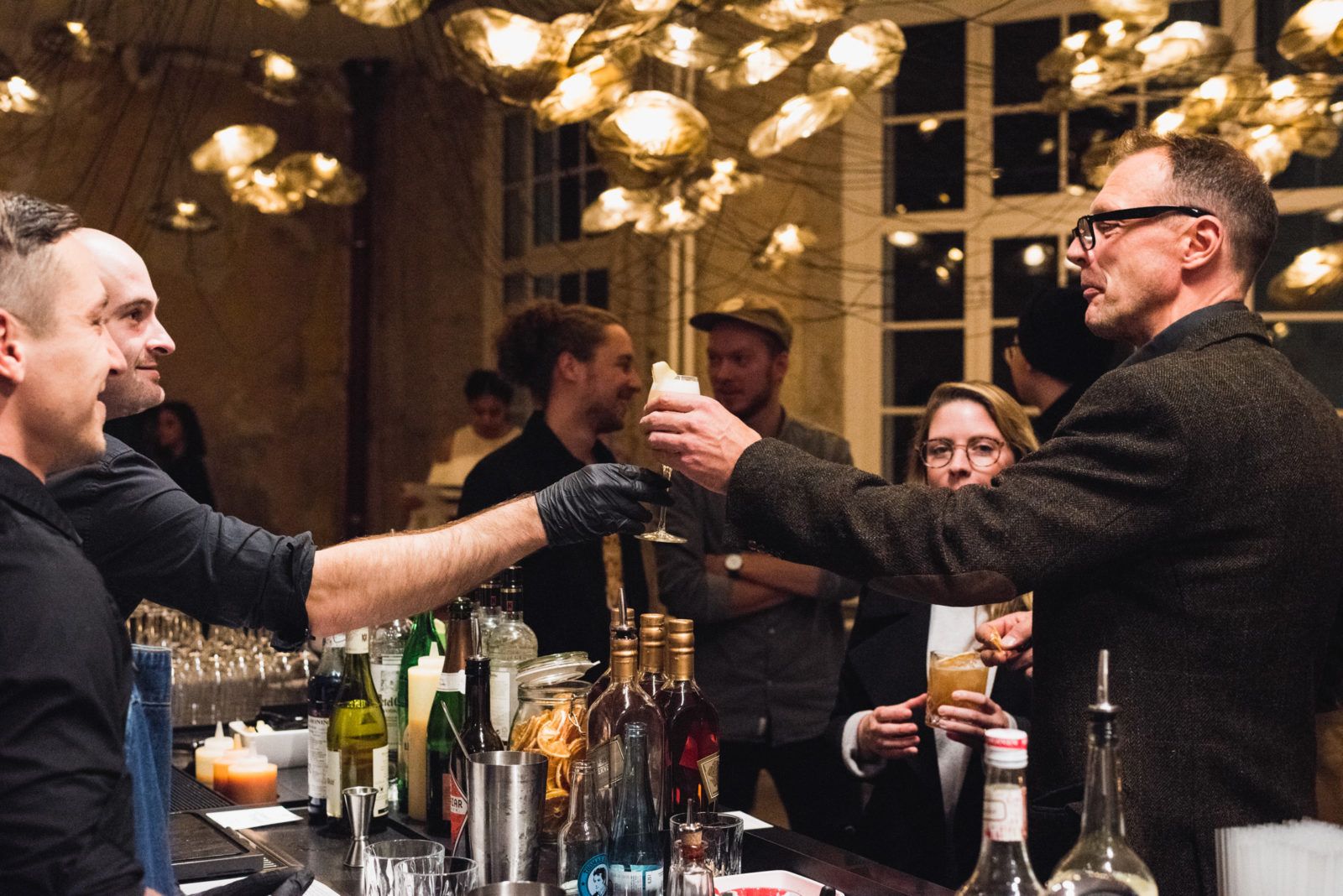 Bartenders serving cocktails under brass pendant lights at a World Class bar night, Friends Space