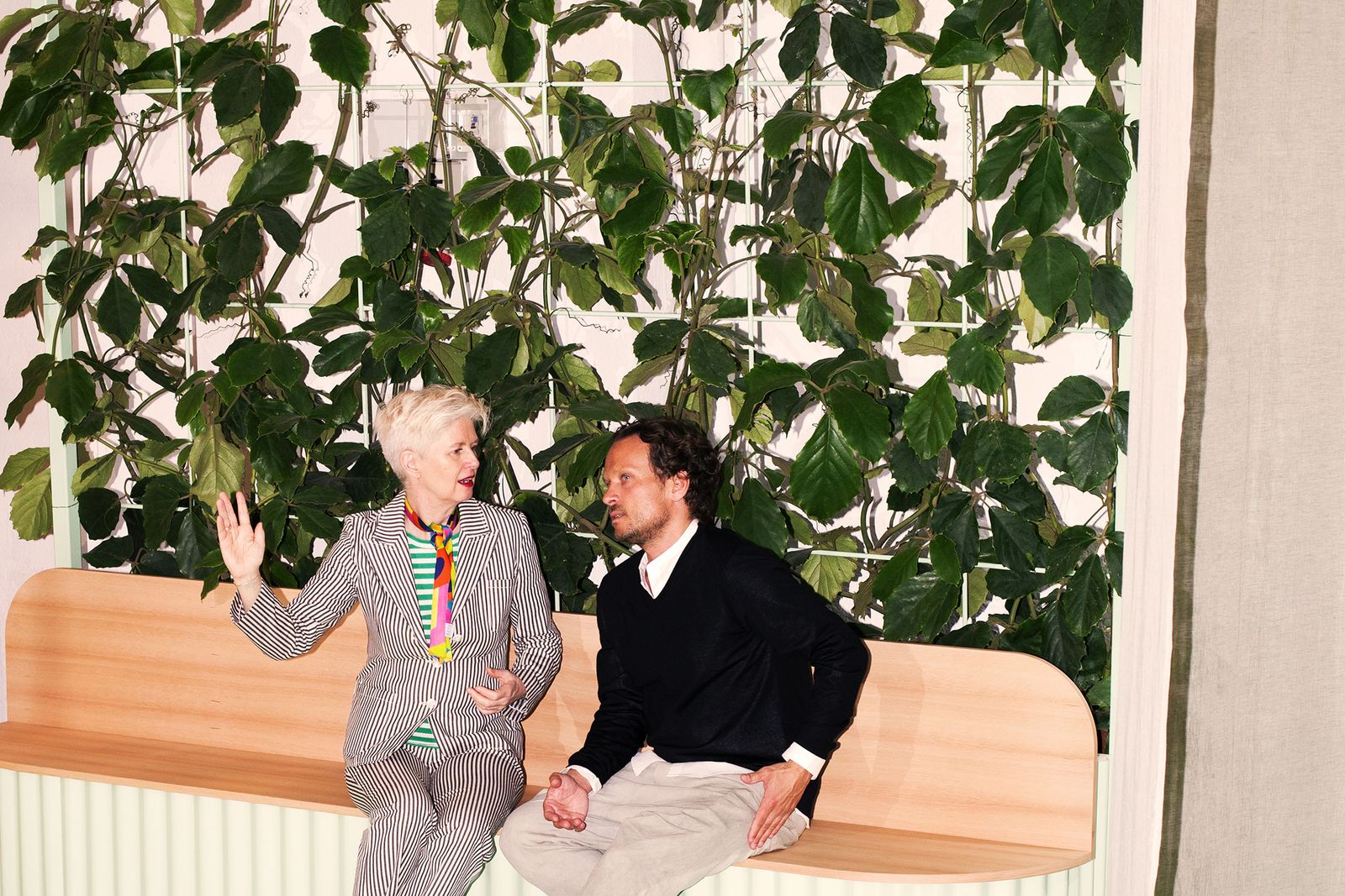 Two people talking on a wooden bench under a green wall at Built by All, Milan