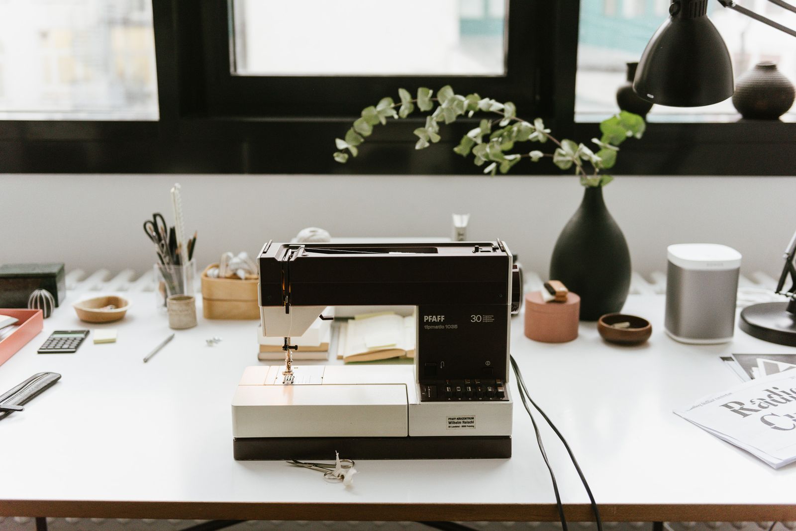 Sewing machine on a white work table with a white Sonos speaker by the window