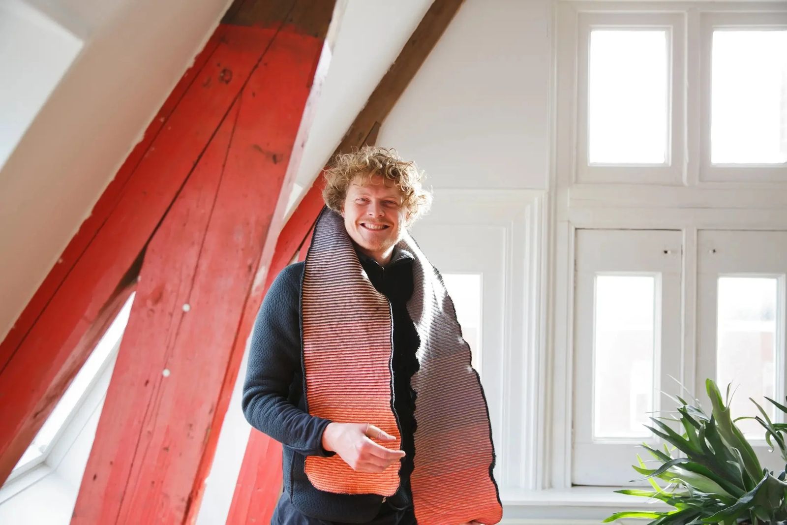BYBORRE founder wearing a knitted textile piece in his studio under red roof beams, Sonos in-home editorial series