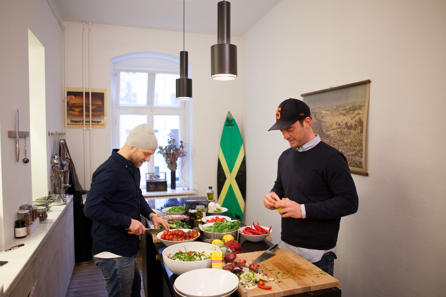 Two friends preparing food in a long Berlin kitchen with a surfboard against the wall