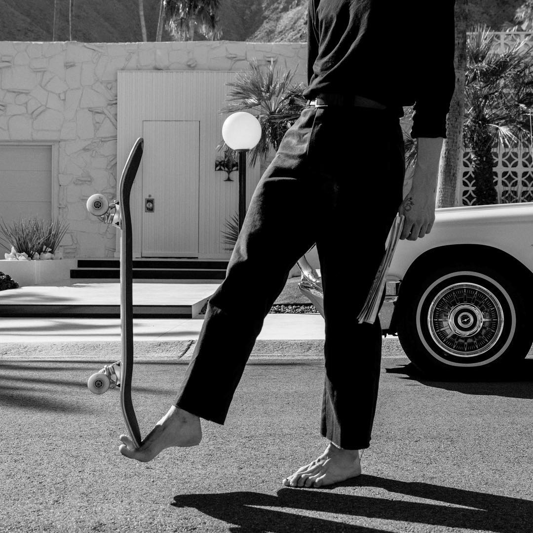 Barefoot on a sunny street, a skateboard balanced on one foot, black and white