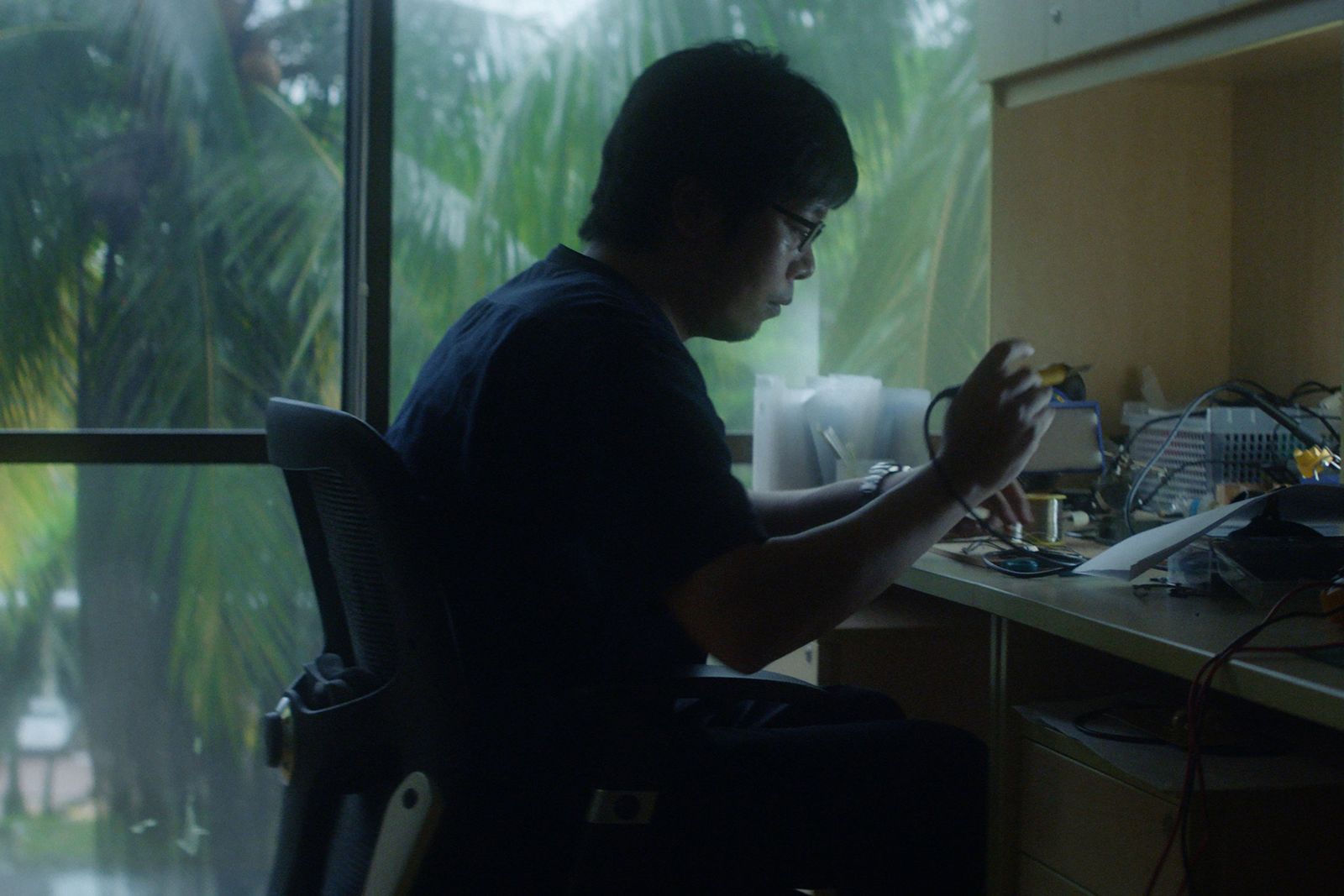 Gadget maker at his workbench by a window with palm trees, Singapore