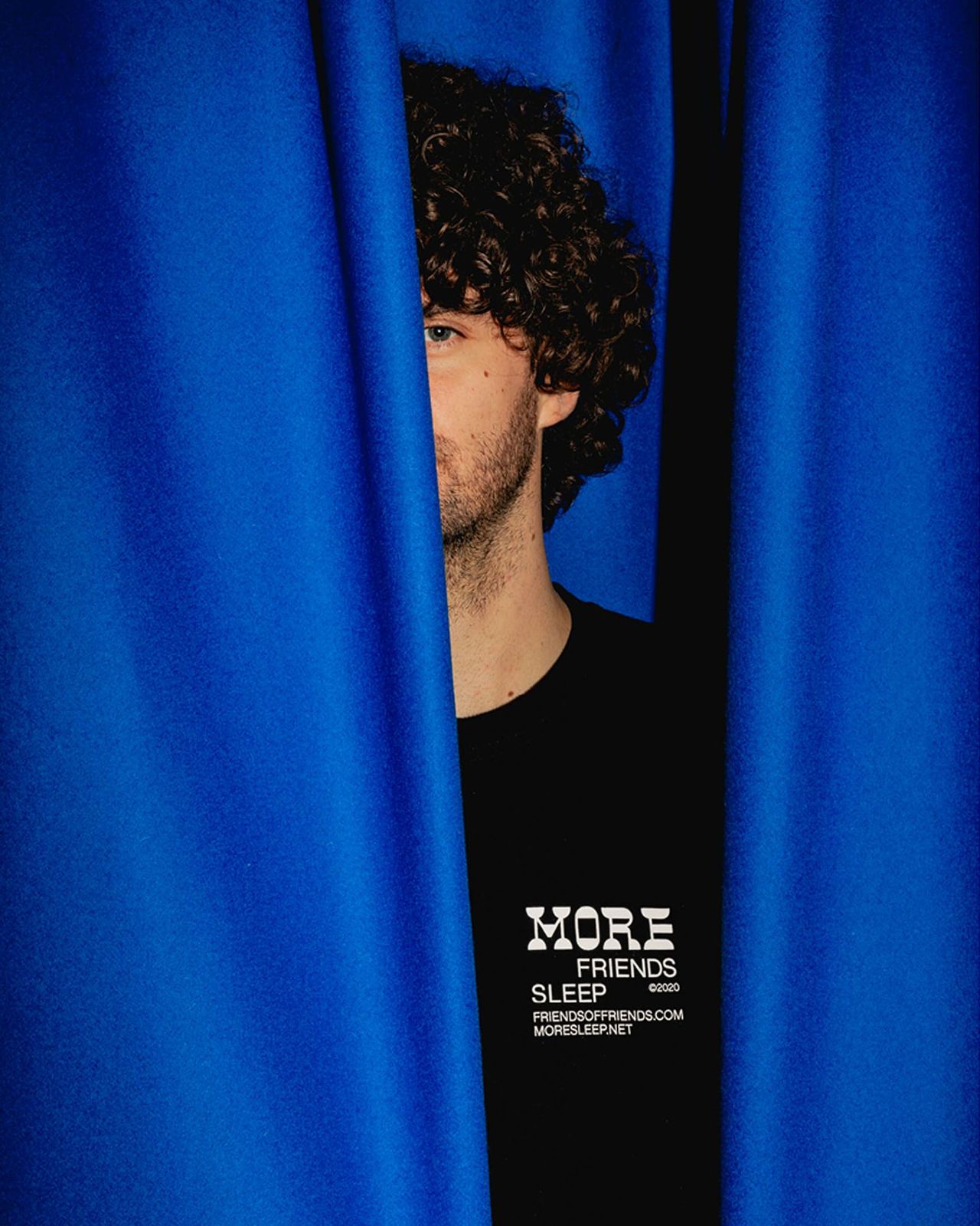 Person in the black MoreFriends T-shirt half hidden behind a blue curtain