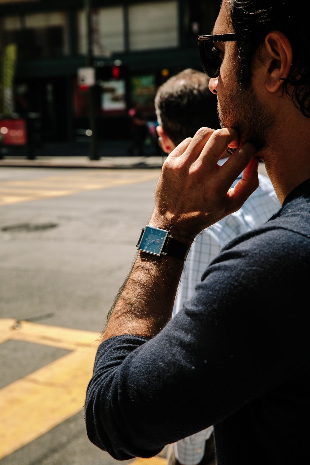 Hand at the chin with a square NOMOS watch, on a sunny street corner