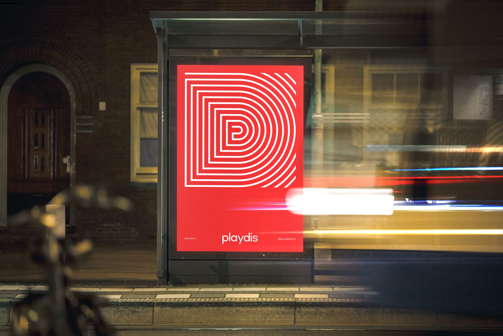Red Playdis poster with the line-drawn P mark in a bus shelter at night, traffic blurring past