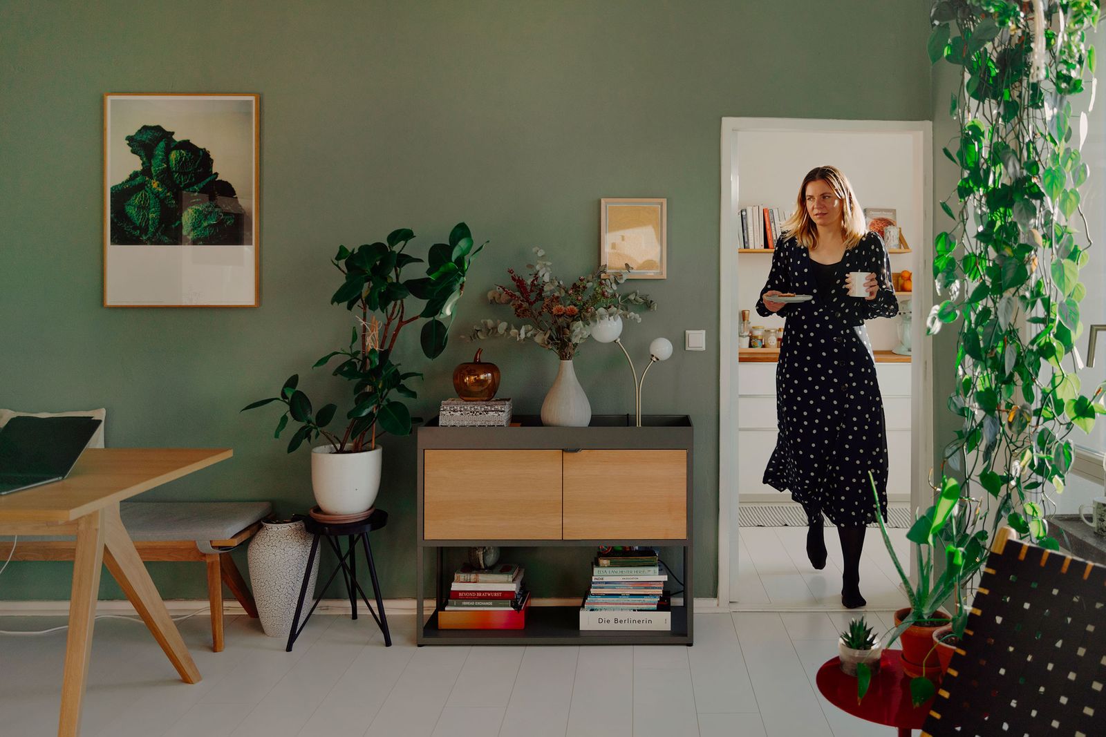 Mary Scherpe walking into a green-walled living room with a coffee cup
