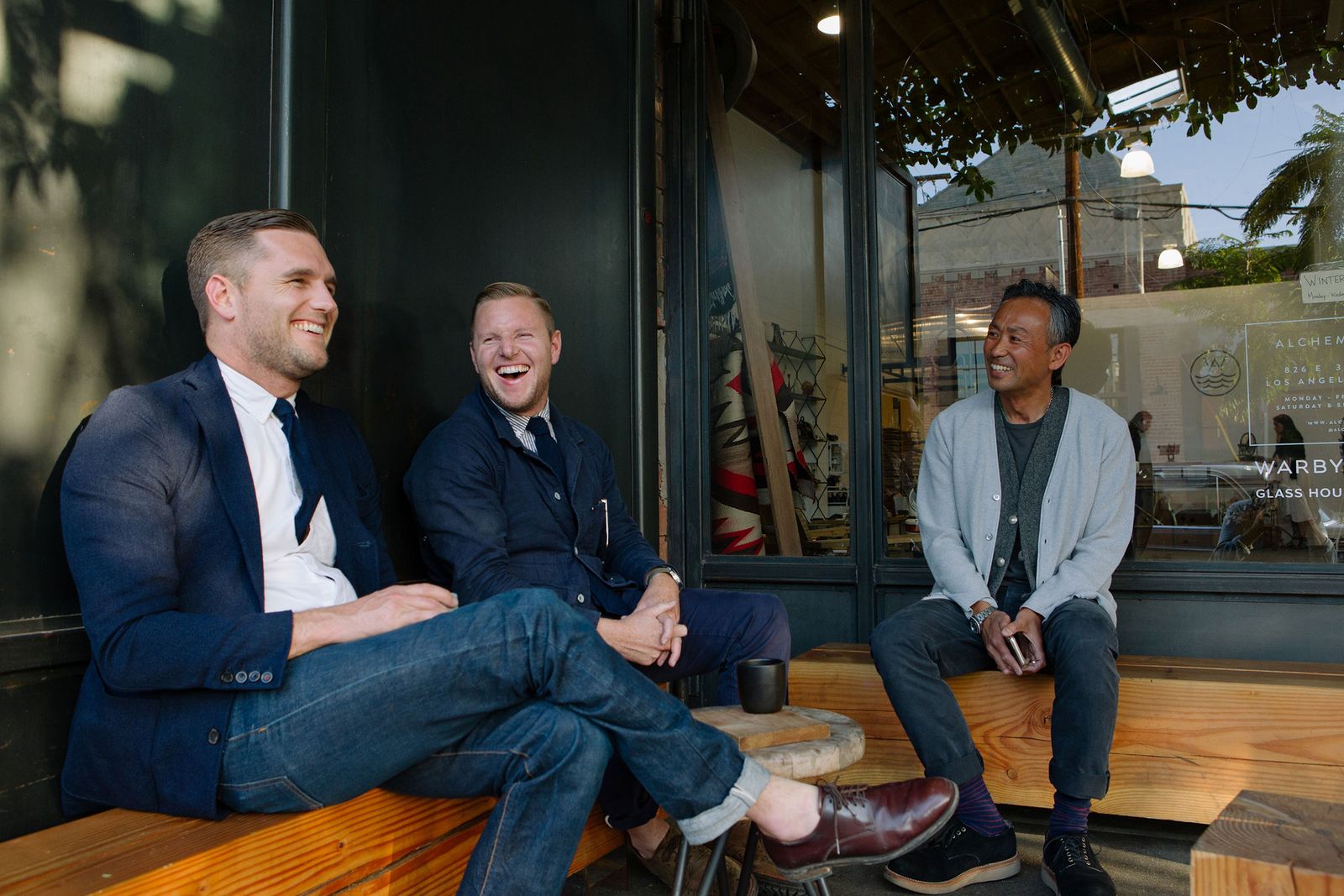 Three men laughing on a wooden bench at a shop front