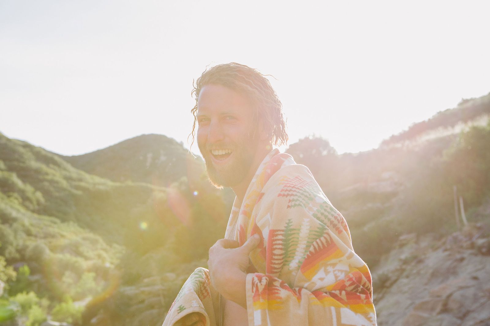 Photographer wrapped in a patterned blanket, laughing in low sun in the hills