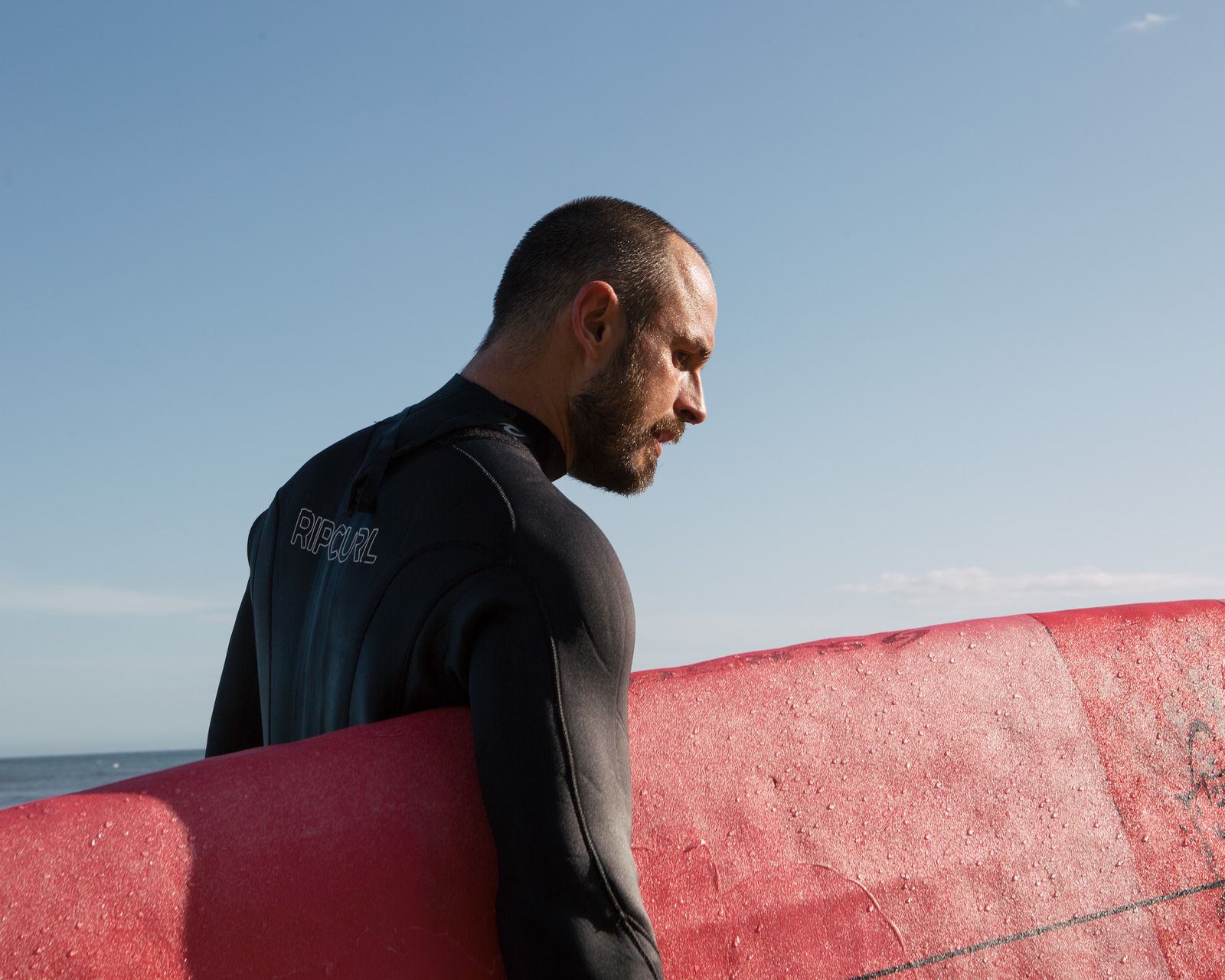 Surfer in a wetsuit carrying a red board at Capitola Beach