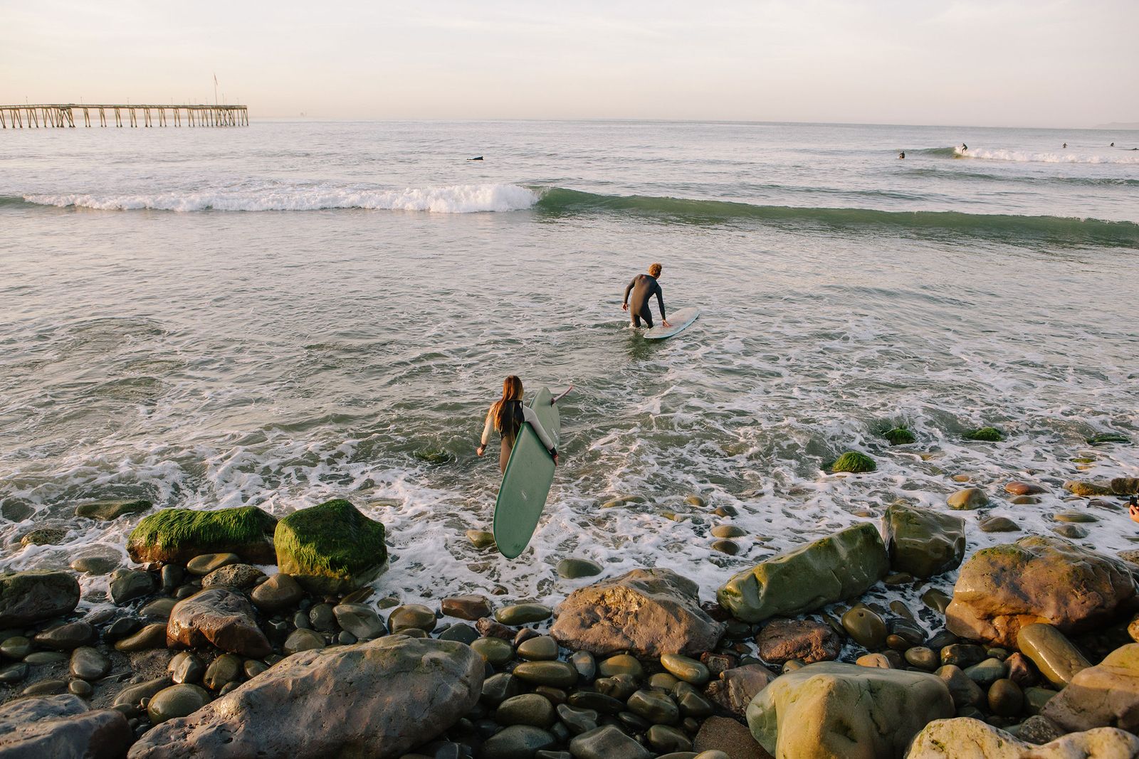 Two surfers wading in over the rocks at Ventura, the pier on the horizon