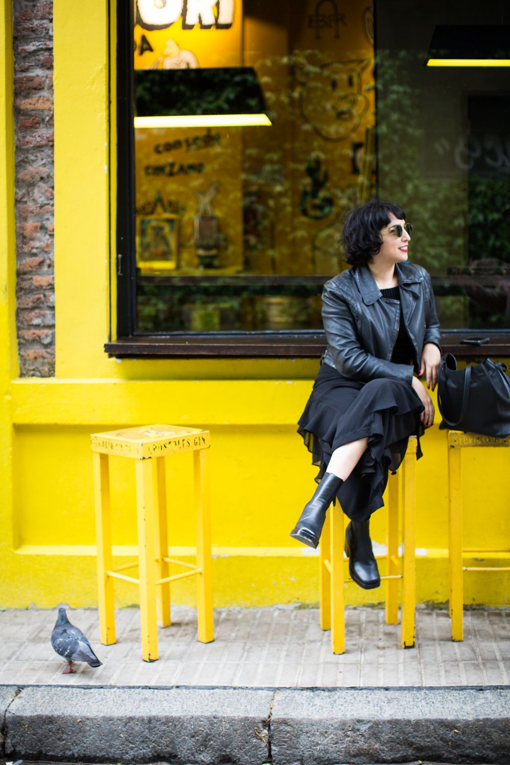 Woman in a leather jacket sitting on a yellow stool outside a yellow café front