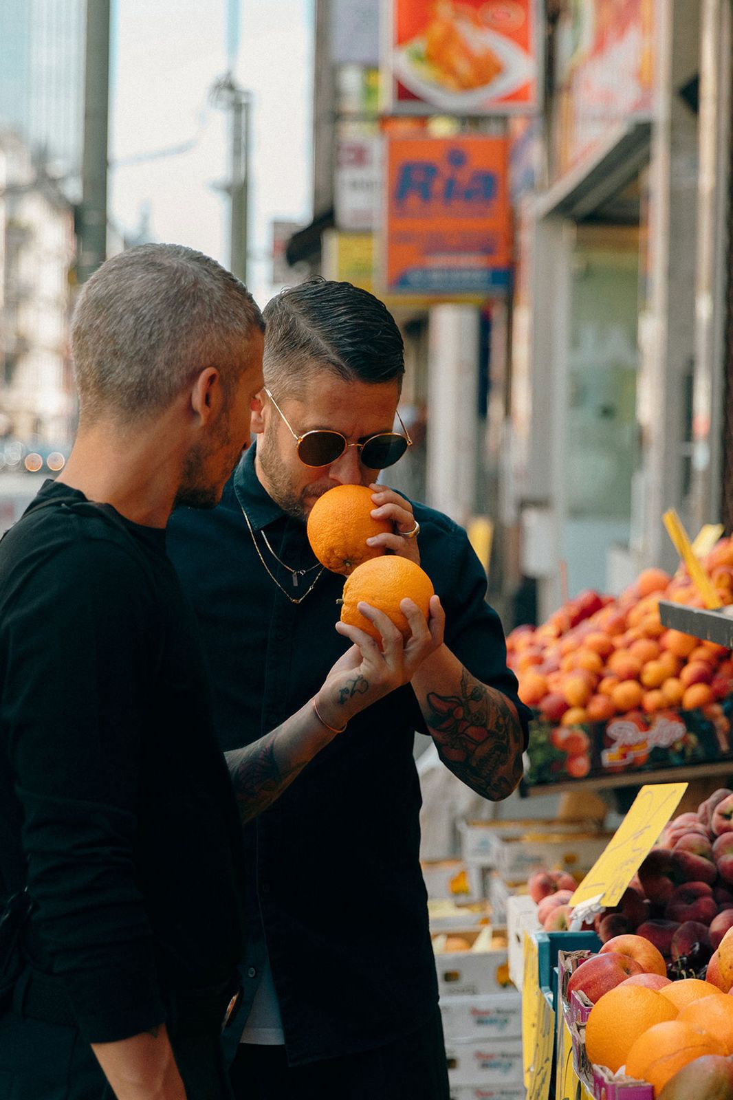 One brother smelling an orange at a fruit stall while the other looks on