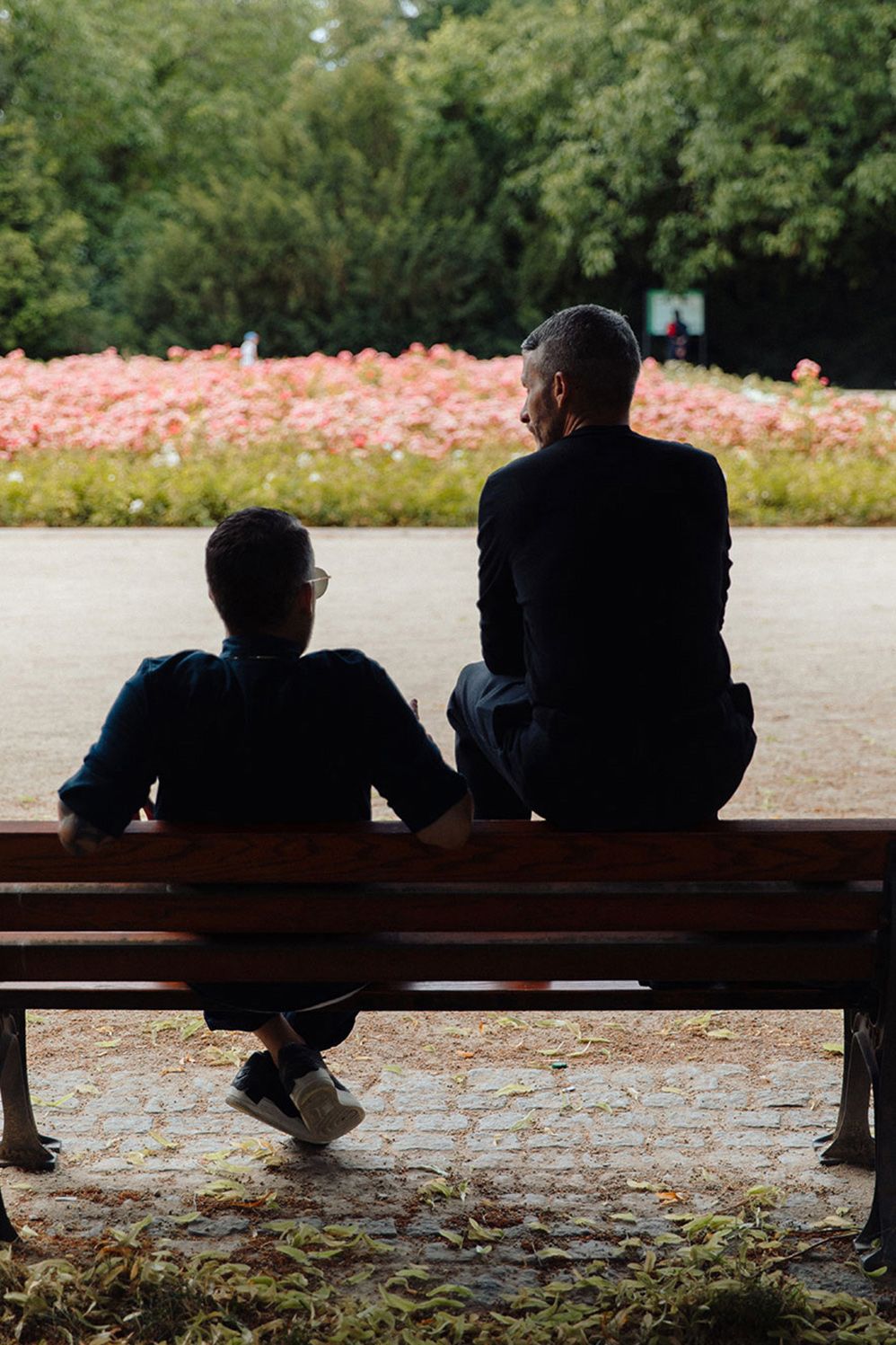 Two men seen from behind on a park bench facing a flower bed