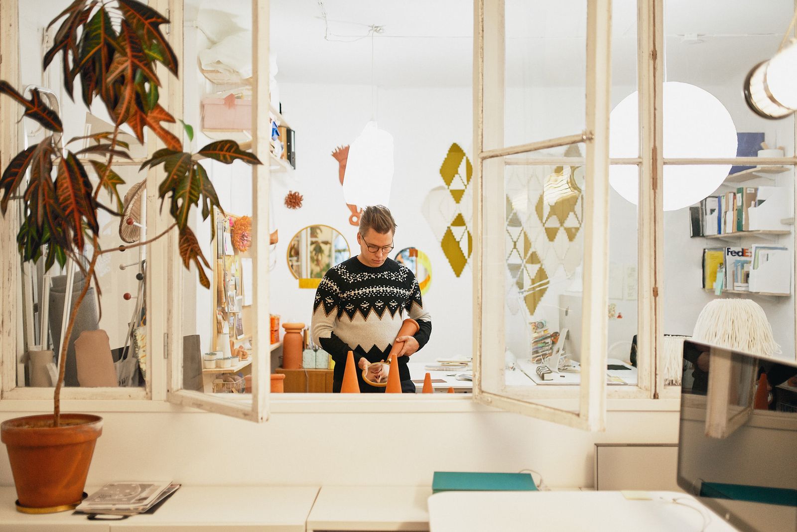 Designer in a patterned knit sweater in a bright Helsinki studio with plants and hanging mirrors